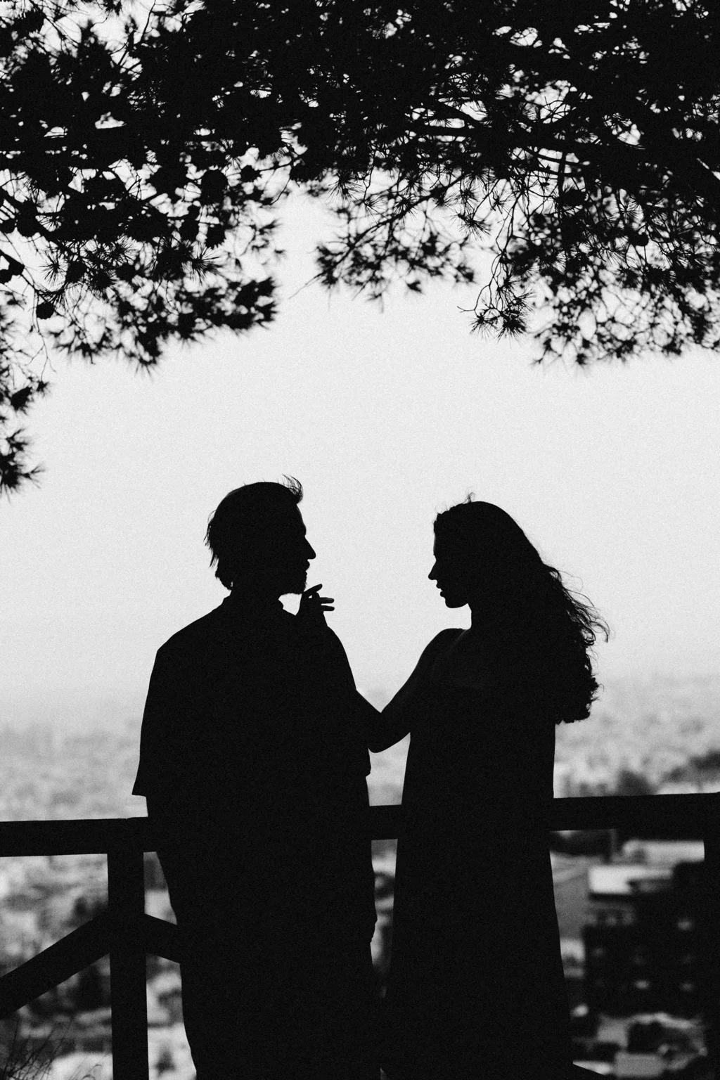 Black and white silhouettes of a couple overlooking Barcelona beneath pine branches